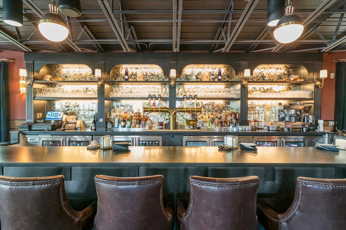 Modern restaurant bar with leather barstools, illuminated shelves of liquor bottles, brass beer taps, and warm ambient lighting.