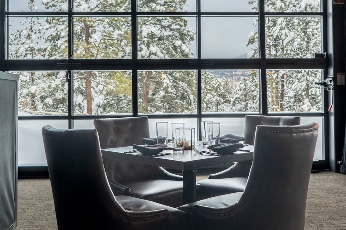 Restaurant dining table set for four beside large windows overlooking snow-covered trees, with upholstered chairs and neatly arranged glassware.