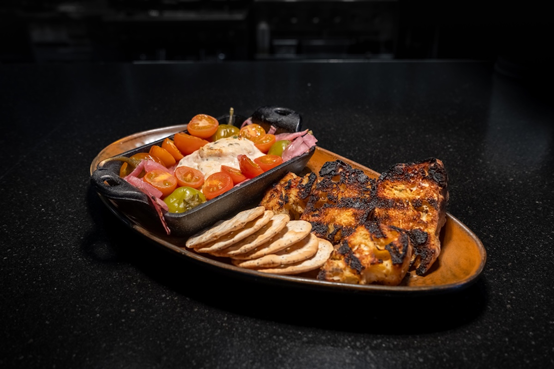 Grilled salmon fillet with charred edges served on a platter with sliced crackers, cherry tomatoes, pickled vegetables, and a creamy dip.
