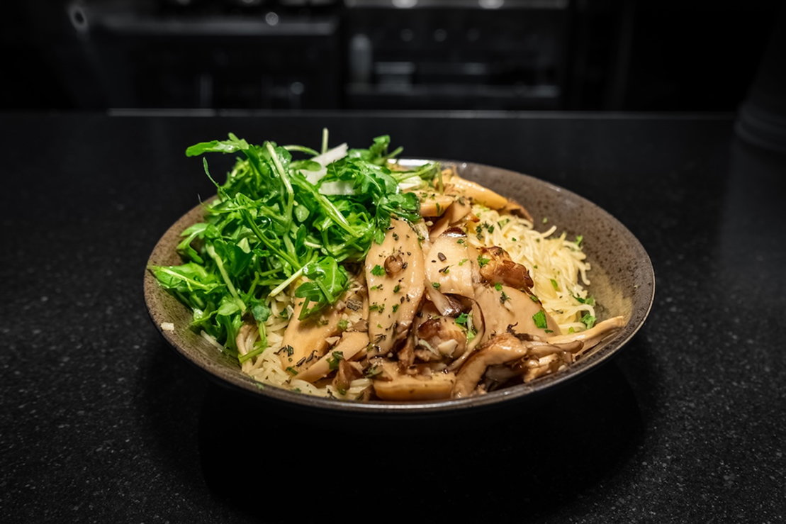 Bowl of pasta topped with sautéed mushrooms, grated cheese, and fresh arugula, served on a dark countertop.