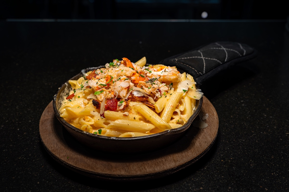 Creamy penne pasta served in a small cast-iron skillet, topped with lobster meat, bacon pieces, and fresh herbs on a dark surface.