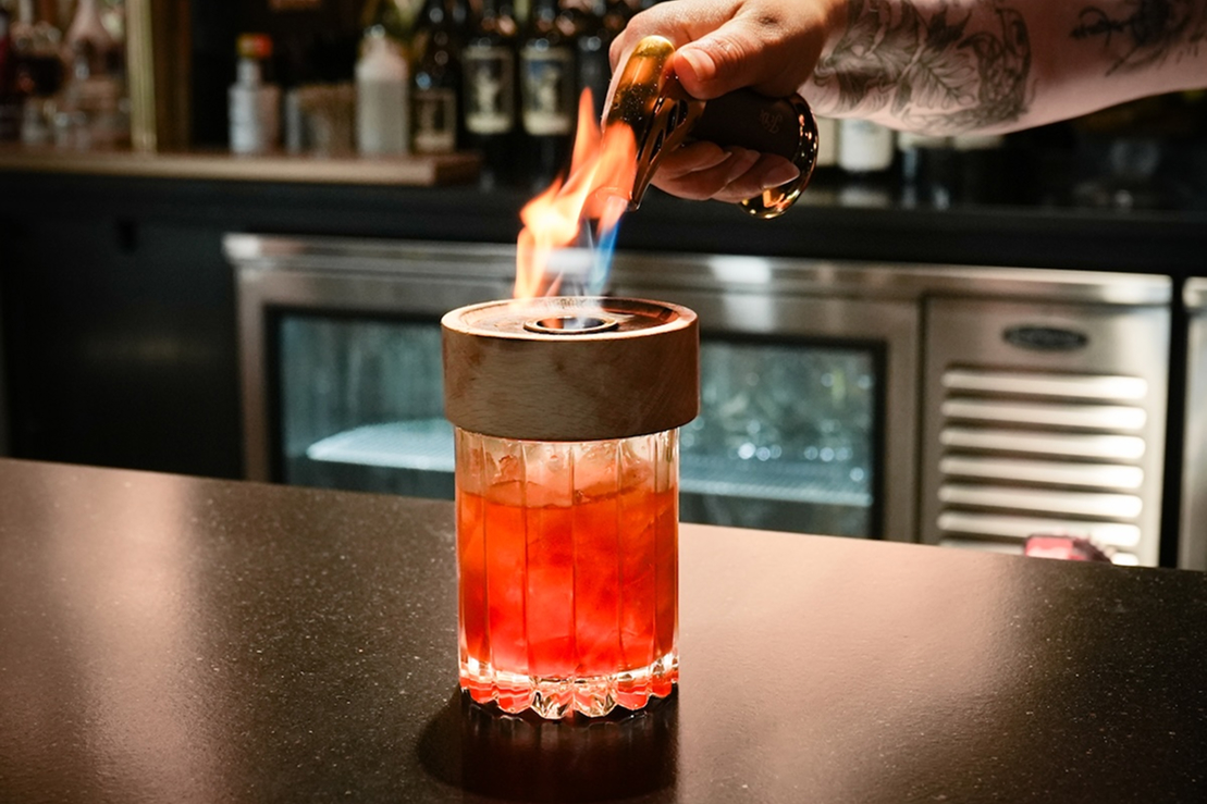 Bartender igniting a cocktail with a flame as a red drink sits on the bar, surrounded by bottles and bar equipment.
