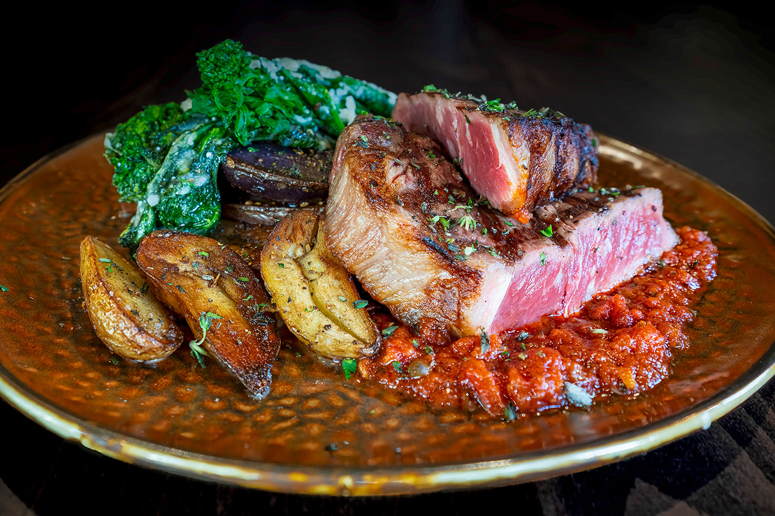 Medium-rare grilled steak sliced and plated with roasted potatoes, sautéed greens, and a red pepper sauce.