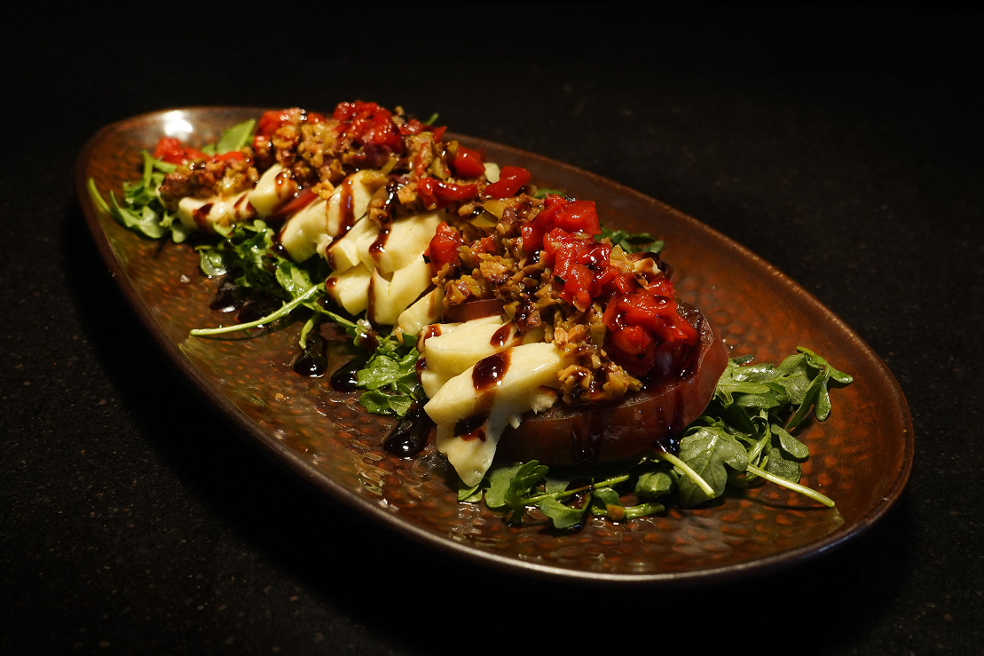 Sliced roasted beet salad topped with melted cheese, chopped vegetables, and balsamic glaze, served over mixed greens on an oval platter.
