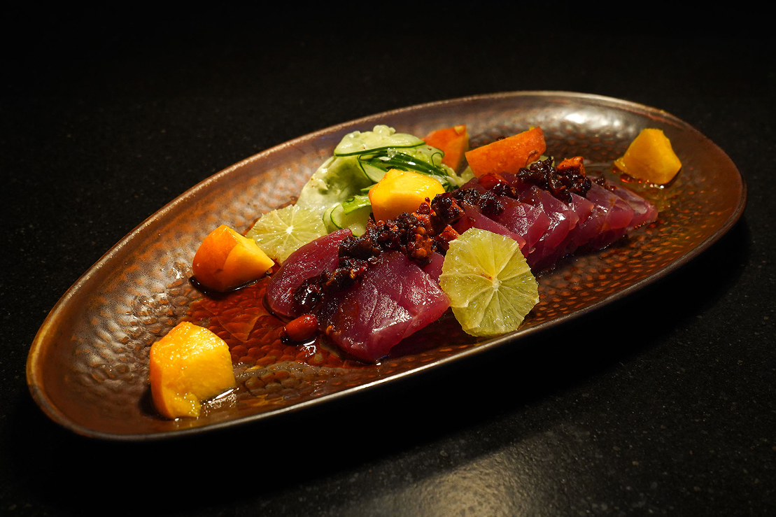 Sliced raw tuna arranged on an oval plate with citrus segments, cucumber, mango, and a dark chili-based topping on a black surface.