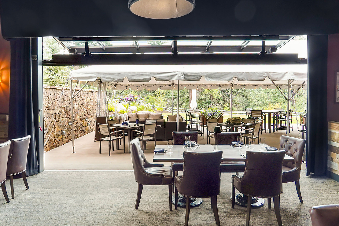 Interior restaurant dining area with tables and leather chairs opening onto a covered outdoor patio with additional seating and garden views.