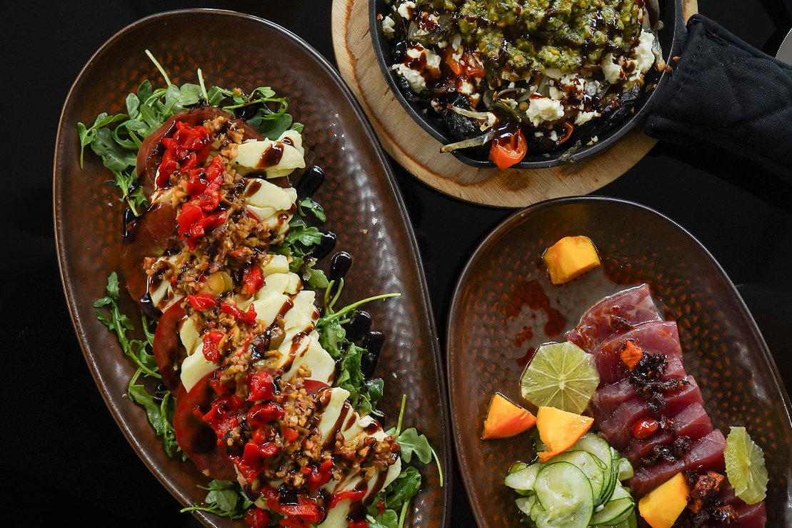 Overhead view of assorted small plates including a caprese-style salad with mozzarella, tomatoes, greens, and balsamic glaze; a baked vegetable dish topped with cheese and pesto; and sliced raw tuna with citrus and cucumber.