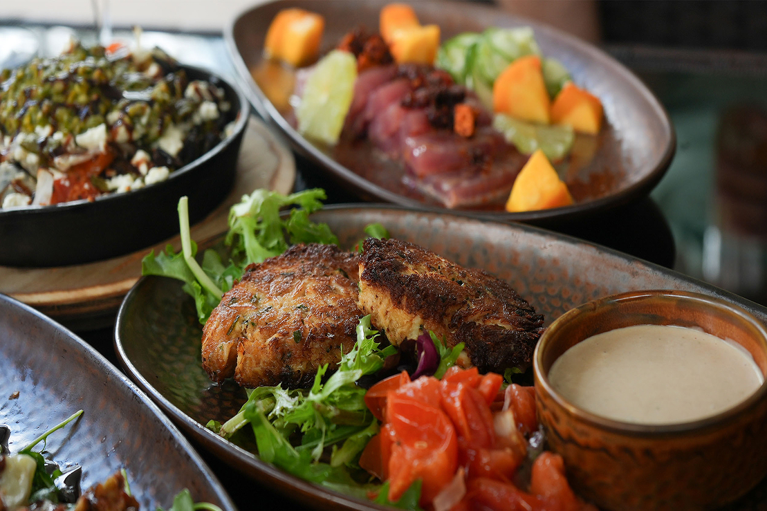 Plated restaurant dishes including pan-seared fish cakes on mixed greens with a creamy sauce, alongside small plates of marinated vegetables and raw tuna with citrus.